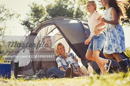 Parents watching happy daughters running around sunny campsite tent