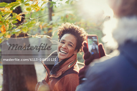 Smiling woman posing for boyfriend with camera phone in sunny autumn woods