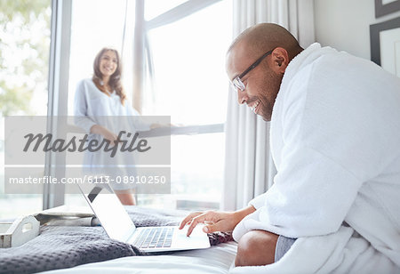 Smiling woman watching boyfriend in bathrobe reading laptop on bed