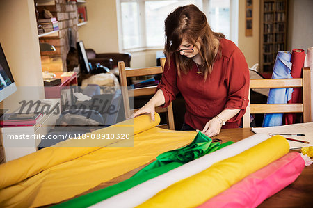 A woman choosing fabric from bolts of brightly coloured material on a tabletop in a workroom.