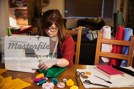 A woman using an electric sewing machine on a table, surrounded by papers and sewing and craft equipment and bolts of cloth.