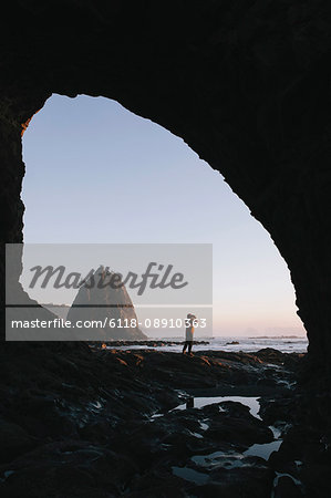 Man standing beneath sea cave at dusk, using binoculars at Hole-in-the-Rock, Rialto Beach, Olympic National Park, Washington, USA.