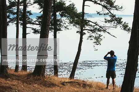 Man standing on a beach, looking through binoculars, San Juan Islands in the distance, Washington, USA.