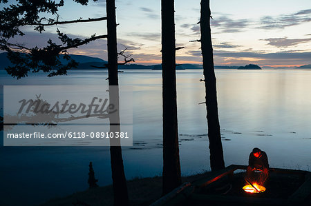 Man sitting by campfire at dusk, San Juan Islands in the distance, Washington, USA. Man sitting by campfire at dusk, San Juan Islands in the distance, Washington, USA.