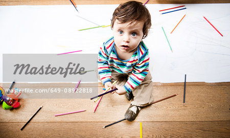 Overhead view of male toddler sitting on floor with coloured pencils and long paper