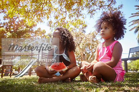 Two young sisters, sitting on grass, eating watermelon