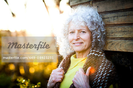 Portrait of mature woman with grey hair leaning against woodland treehouse at sunset
