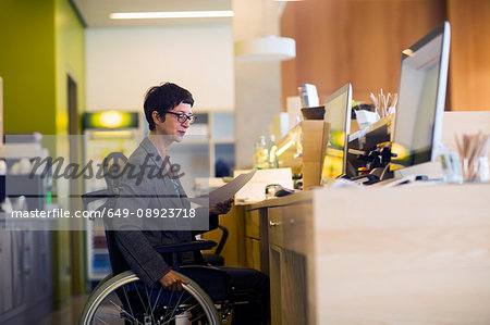 Woman in wheelchair, sitting at desk, looking at document