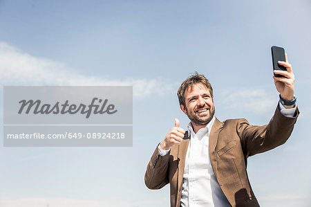 Businessman giving thumbs up for smartphone selfie against blue sky