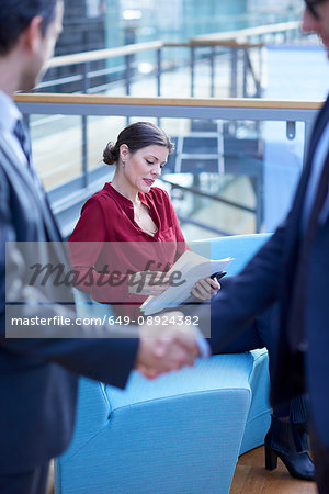 Businessmen shaking hands looking at businesswoman reading paperwork in office atrium