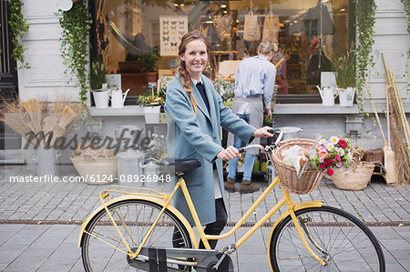 Portrait smiling woman walking bicycle with flowers in basket outside storefront