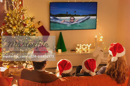 Family in Santa hats watching summer holiday video on TV in Christmas living room