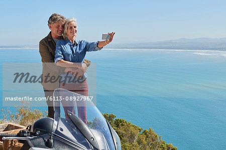 Senior couple taking selfie next to motorcycle overlooking sunny ocean view