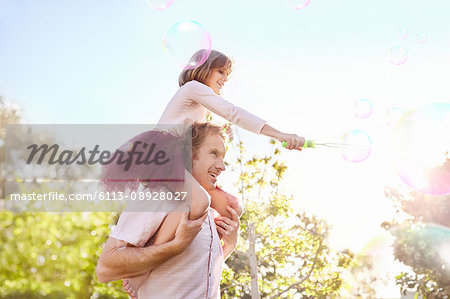 Father carrying daughter with bubble wand on shoulders in sunny summer park