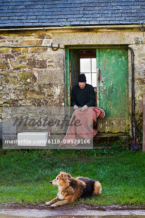 Man sitting in the doorway of a sailmaker's workshop, sewing a sail, a dog lying on the grass in the foreground.