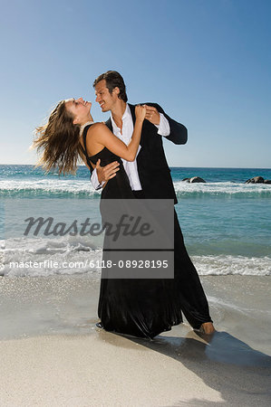 man in suit dancing with blond woman in black evening gown on a sandy beach.