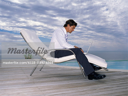 A man in a white shirt and black trousers in a sun lounger chair with a laptop on his lap.