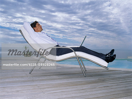 A man in a white shirt and black trousers in a sun lounger chair with a laptop on his lap.