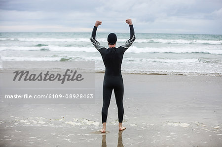 Rear view of athlete in wet suit standing with arms up on beach