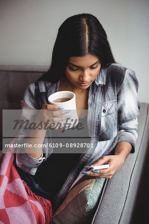 Woman using mobile phone while holding cup of coffee in living room at home