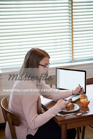 Woman using mobile phone while having breakfast in living room at home