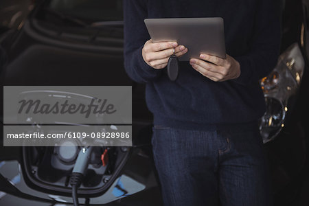Mid section of man using digital tablet while charging electric car in garage