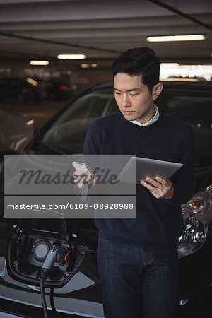 Man using digital tablet and mobile phone while charging electric car in garage