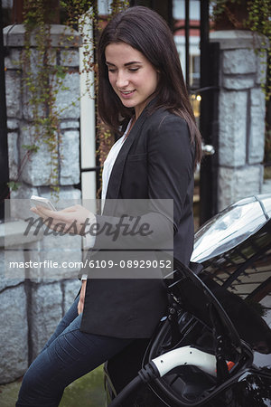 Beautiful woman using mobile phone while charging electric car at vehicle charging station