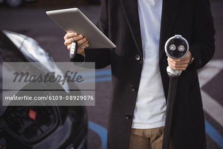 Mid section of man using digital tablet while holding car charger at electric vehicle charging station