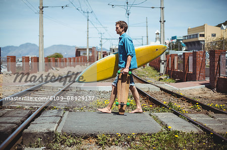 Man carrying skateboard and surfboard crossing railway track