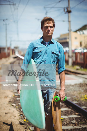 Portrait of man carrying skateboard and surfboard crossing railway track