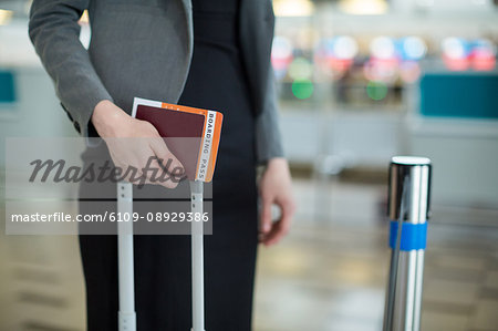 Mid section of businesswoman standing with boarding pass, passport and luggage at airport terminal