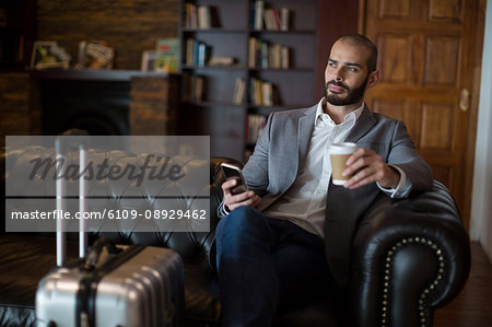 Thoughtful businessman holding mobile phone and coffee cup in waiting area at airport terminal