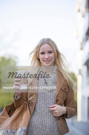 Pretty blonde woman with cafe to go walking in city center and smiling at camera
