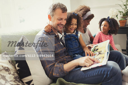 Smiling multi-ethnic father reading book with daughter on sofa