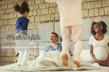 Multi-ethnic pregnant parents watching daughters jumping on bed Multi-ethnic pregnant parents watching daughters jumping on bed