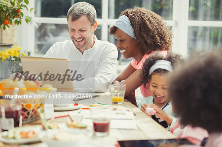 Smiling young multi-ethnic family using laptop and eating breakfast at table