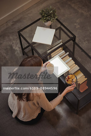 Woman with digital tablet having breakfast in living room at home