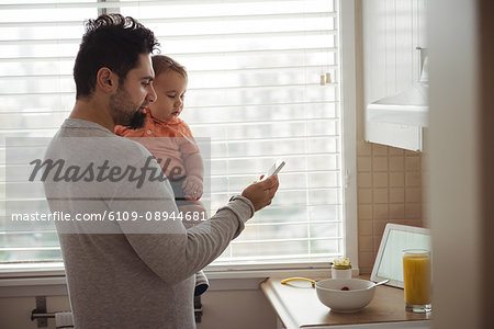 Father using mobile phone while holding his baby in kitchen at home