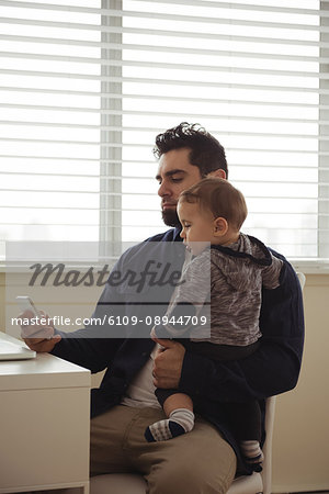 Father holding his baby while using mobile phone at desk at home