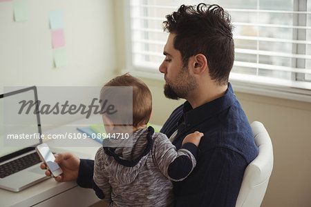 Father holding his baby while using mobile phone at desk in home