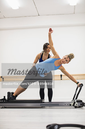Female trainer assisting woman with stretching exercise on reformer in gym