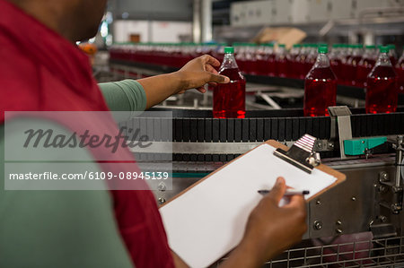Mid section of male worker inspecting red juice bottles on production line in factory