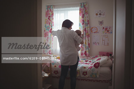 Rear view of mother with baby standing by window in bedroom