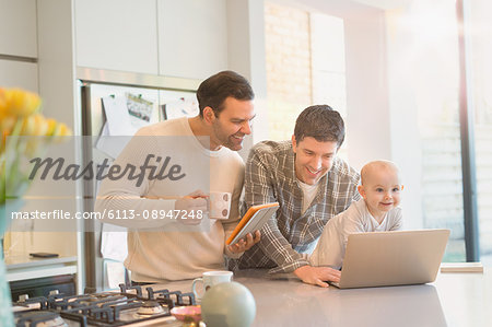 Male gay parents with baby son using digital tablet and laptop in kitchen