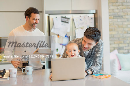 Male gay parents and baby son using laptop and digital tablet in kitchen