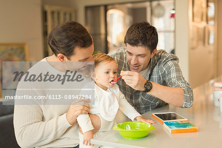 Male gay parents feeding baby son in kitchen Male gay parents feeding baby son in kitchen