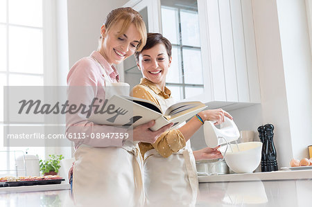 Female caterers with cookbook baking in kitchen, using electric hand mixer