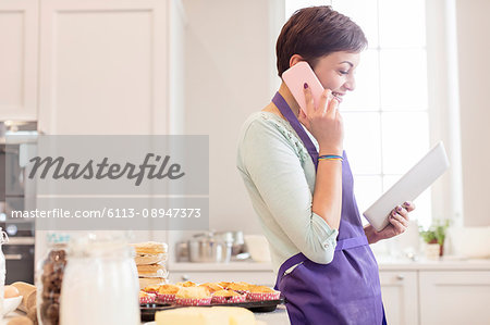 Female caterer baking, talking on cell phone and using digital tablet in kitchen