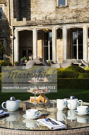 A table in a garden outside an old house, with cake stand and a selection of cakes and sandwiches set for a traditional afternoon tea.